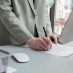 Unrecognizable worker standing at table with computer while taking notes in document while working in office